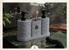 A gray three-compartment bathroom organizer with embossed geometric patterns, black pump dispensers, a wooden toothbrush, and a metal spoon, set on a dark marble countertop in a bathroom.