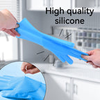 A pair of light blue silicone cleaning gloves with textured scrubbing sides, being held by two hands. The background is a blurred kitchen setting, and an inset circle shows a close-up of the scrubbing texture.